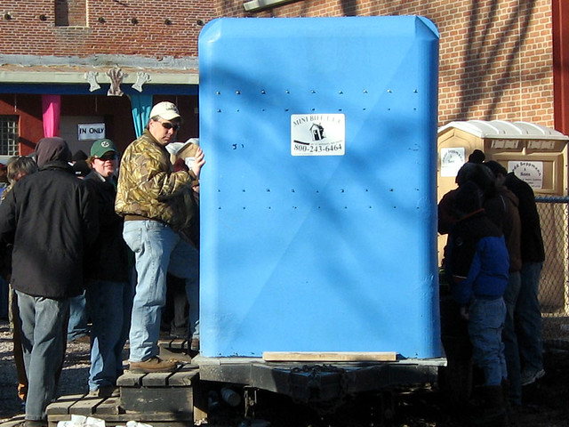 A bunch of guys waiting in line for the urinal trailor while 4 guys pee on the back of it.