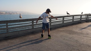 Some dude glidin down the ocean beach pier on some little skate platform things